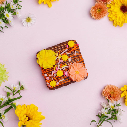 a square brown cookie with yellow and orange frosting and sprinkles surrounded by flowers