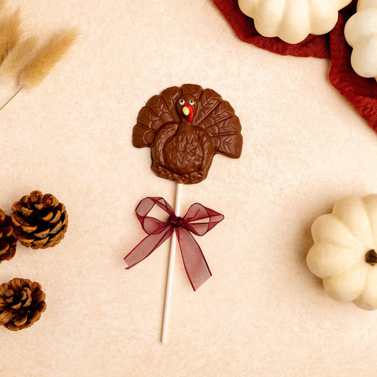 a chocolate turkey lollipop next to white pumpkins and pine cones