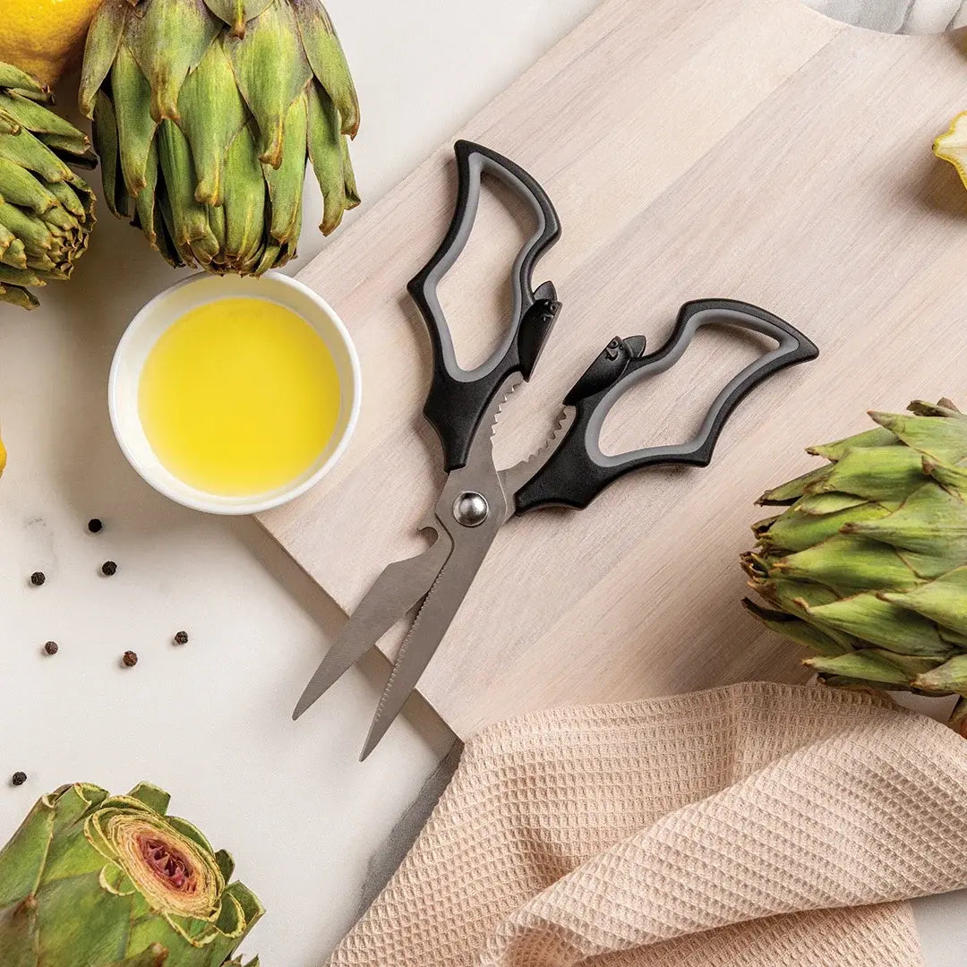a cutting board with scissors and a bowl of oil next to artichokes