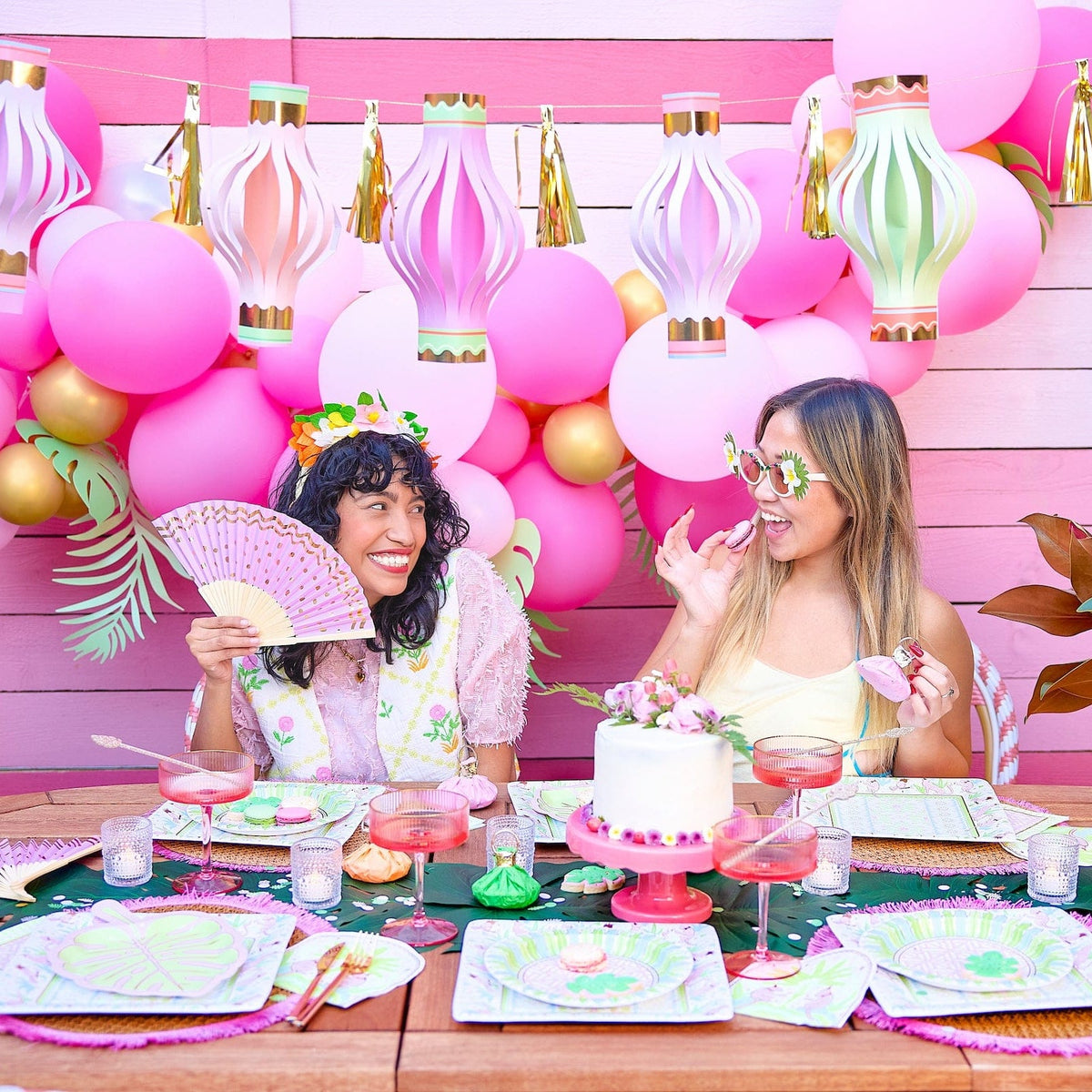 two women sitting at a table with pink balloons and paper fans