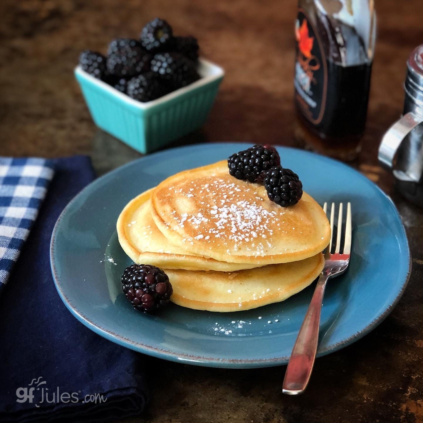 Stack of pancakes with blackberries on a blue plate with syrup and berries in the background.