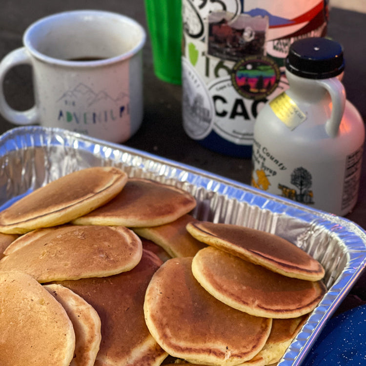 Stack of pancakes in a metal tray with a coffee mug, bottles, and cups in the background.
